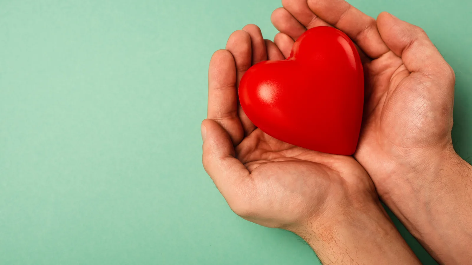 Two hands cupped together holding a red heart against a green background.