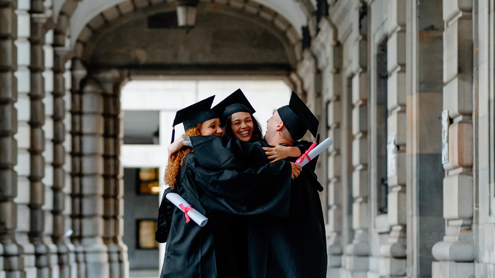 Graduates in caps and gowns embracing in a university colonnade.