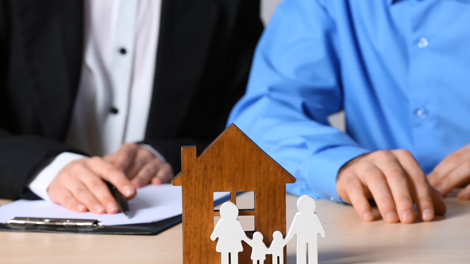 Two people at a table beside a small wooden house and cut-out family figures.