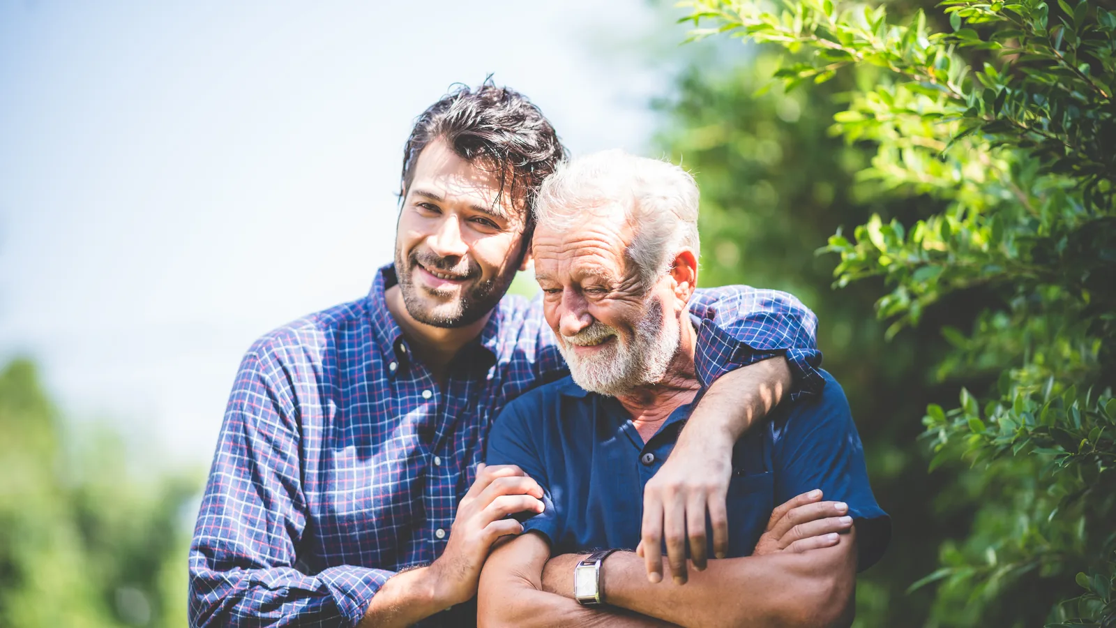 An adult son with his arm around his older father, both smiling, outdoors.