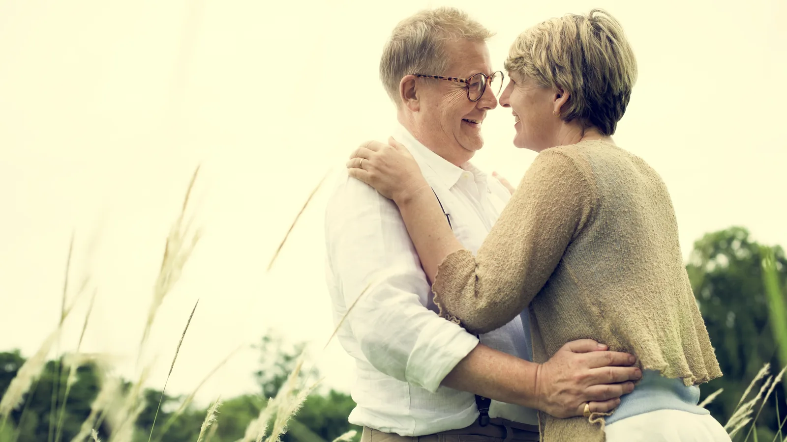 An older couple embracing outdoors among tall grass, smiling at each other.