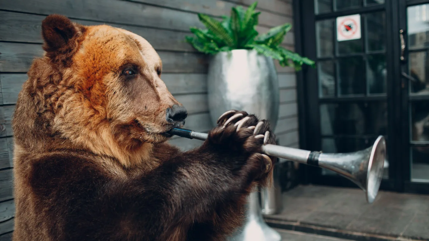 A brown bear standing upright, holding and playing a trumpet outside a wooden building.