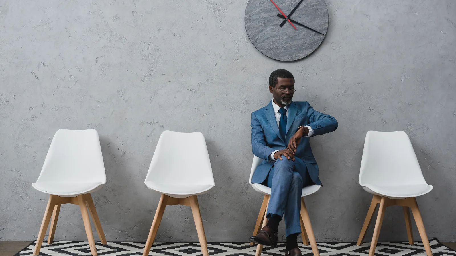 A man sitting alone in a row of empty chairs beneath a wall clock.