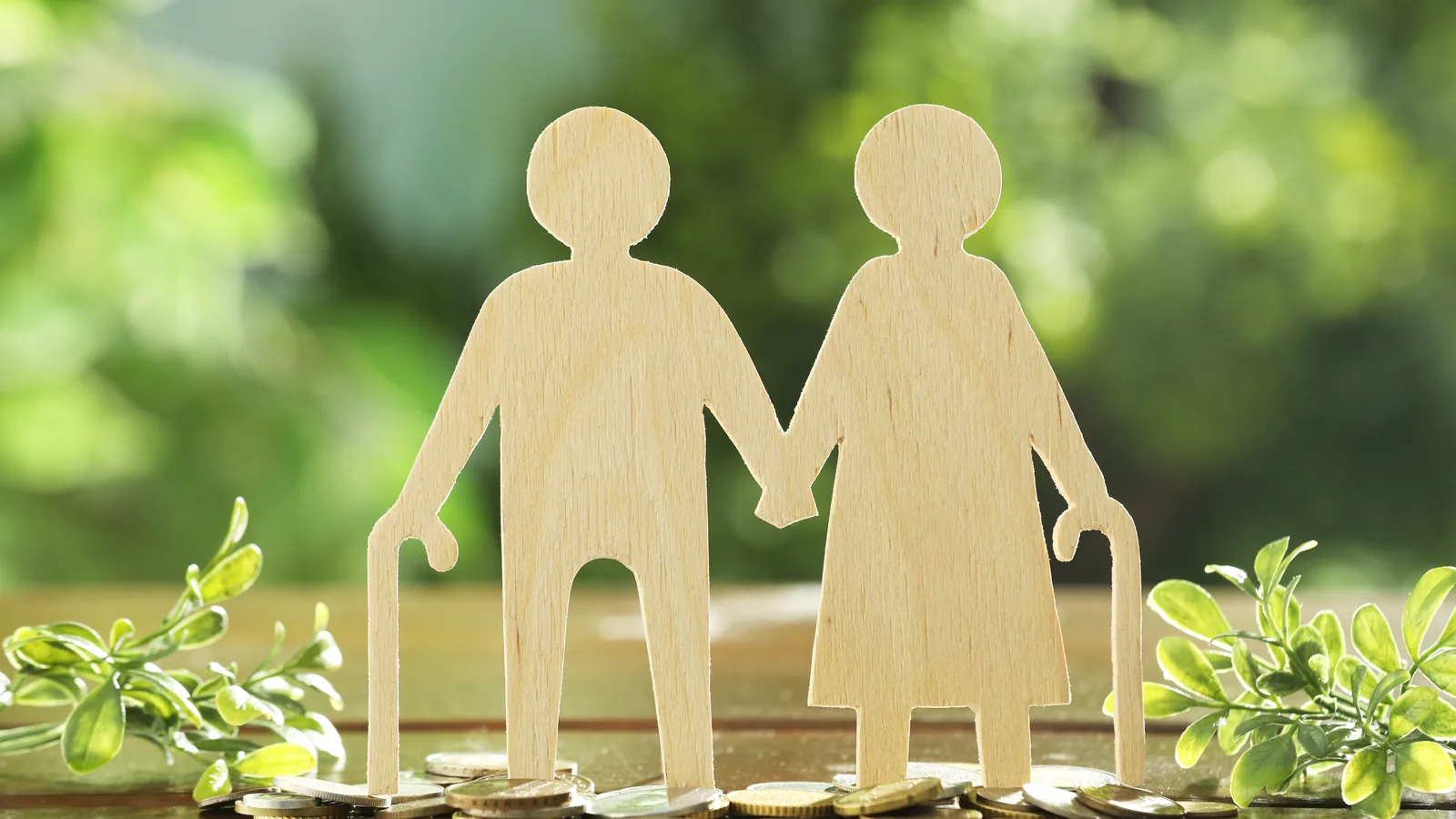 Two wooden figures of an older couple holding hands among greenery.