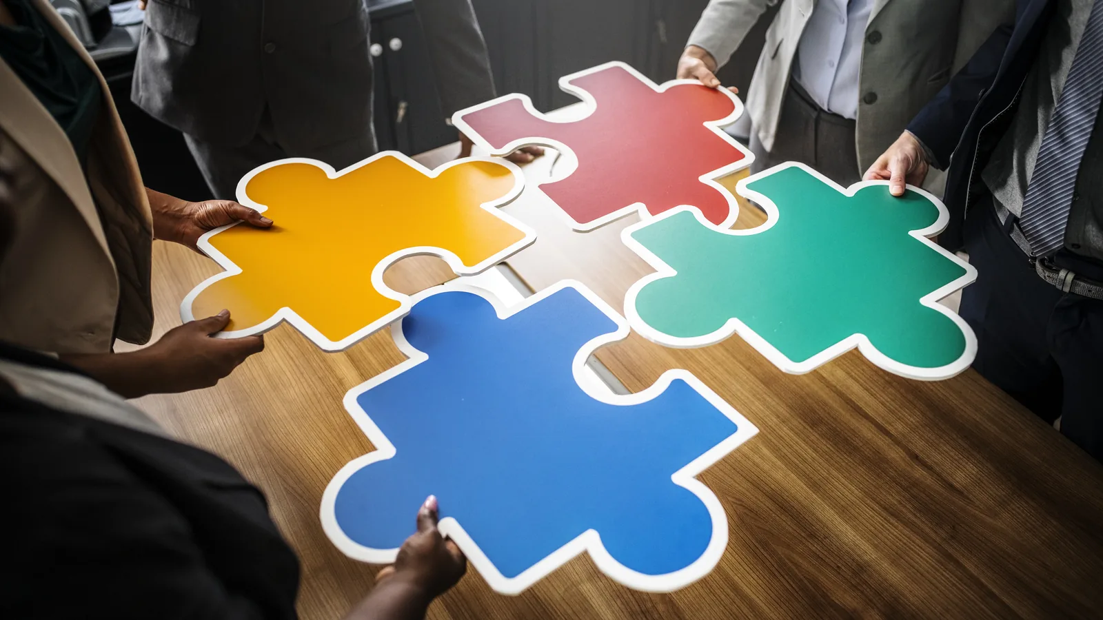 Several hands fitting large colored puzzle pieces together on a table.